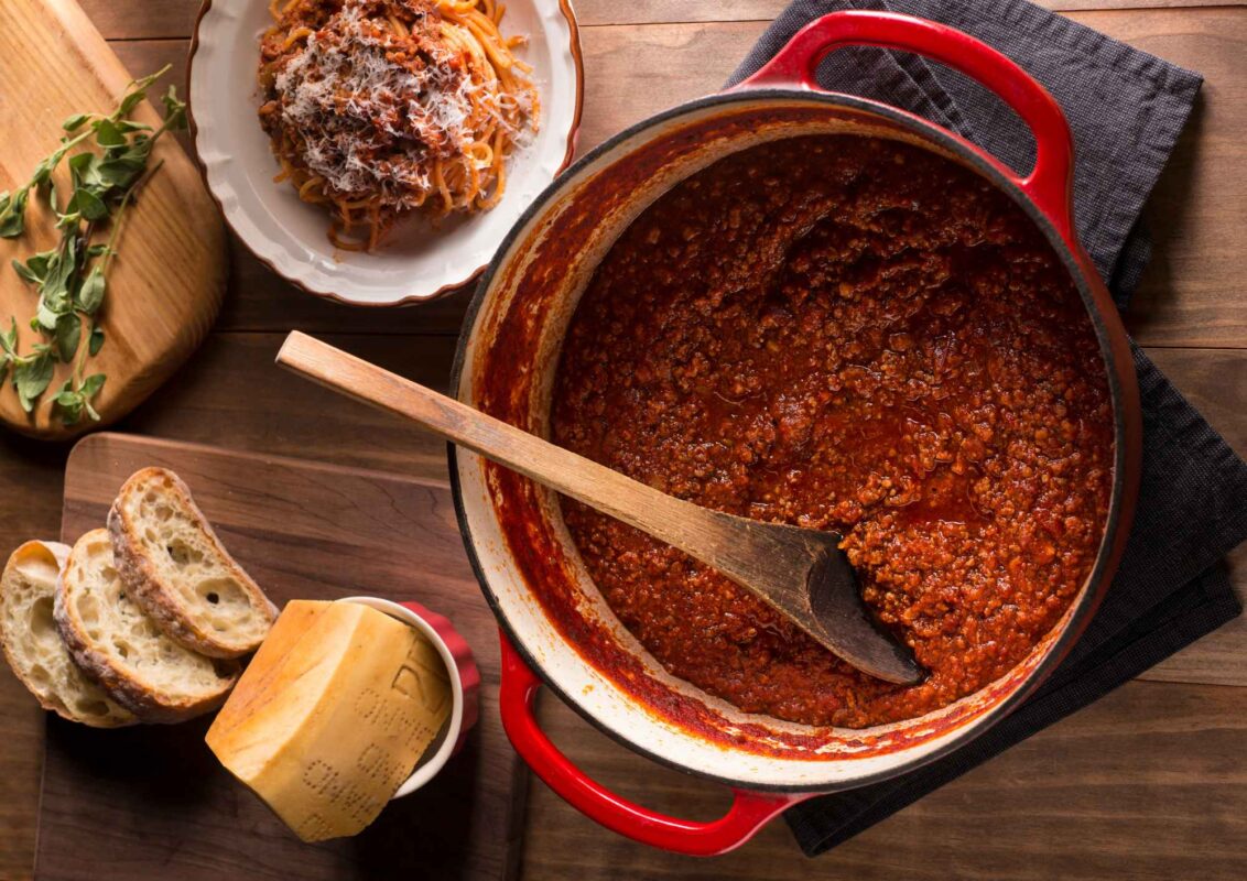 Bolognese wird vorbereitet in einem Topf, daneben ein fertig angerichteter Teller, Parmesan, Brot und frische Kräuter